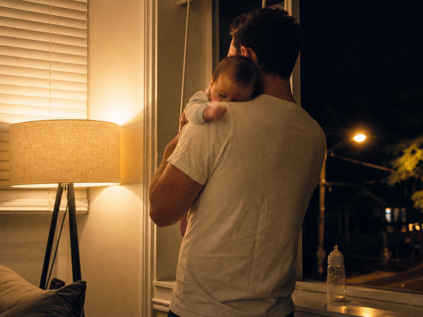 A father at the dark window in the middle of the night, baby asleep on his shoulder.